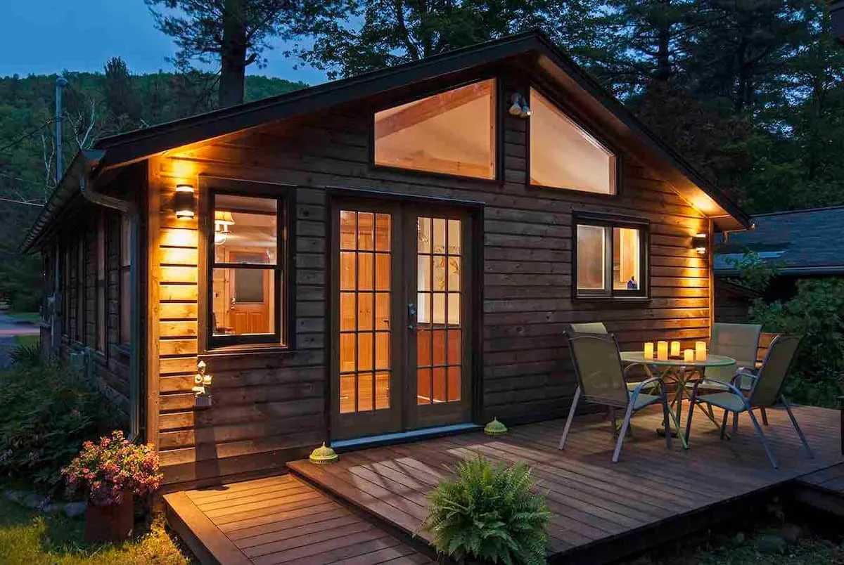 Timber cabin at dusk, windows lit warm, chairs on the deck.